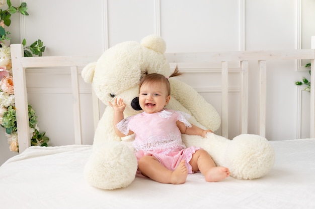 Bonne petite fille de six mois assise sur un lit blanc dans des vêtements roses, avec un gros ours en peluche et souriant