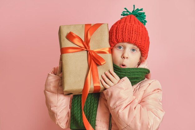 Bonne petite fille souriante caucasienne mignonne avec boîte-cadeau de Noël avec ruban rouge. fille dans une écharpe tricotée et un chapeau sur fond rose, espace de copie de concept d'humeur de noël