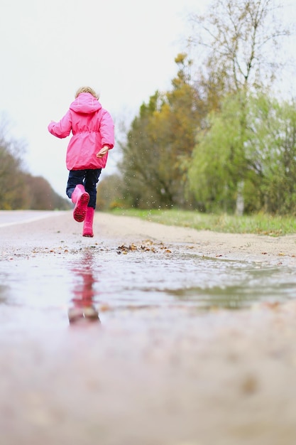 Bonne petite fille en veste imperméable rose bottes en caoutchouc saute joyeusement à travers les flaques d'eau sur la route par temps de pluie Printemps automne Amusement des enfants à l'air frais après la pluie Loisirs en plein air