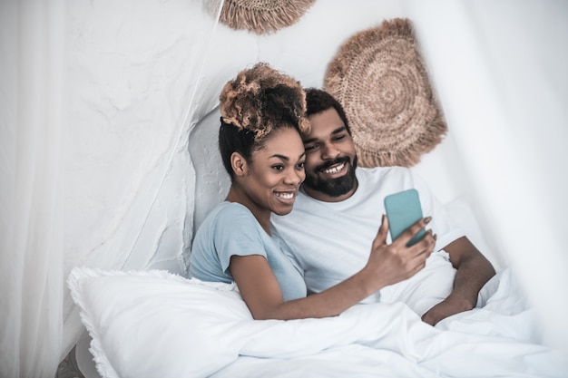 Bonne photo. Jeune femme et homme à la peau sombre prenant selfie dans son lit à joyeusement à l'écran du smartphone