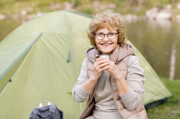 Bonne randonneuse mature avec boisson chaude vous regarde tout en profitant du repos sous la tente après un long voyage dans la forêt