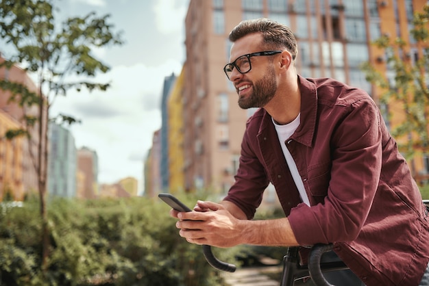 Bonnes nouvelles. Vue latérale d'un bel homme avec du chaume dans des vêtements décontractés et des lunettes se penchant sur son vélo, tenant un téléphone portable et souriant tout en se tenant à l'extérieur. Notion numérique. Activité du week-end