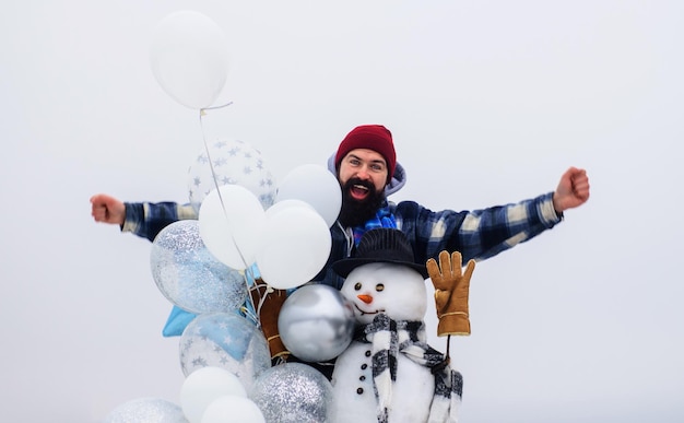 Bonnes vacances homme barbu en vêtements d'hiver avec bonhomme de neige en bonnet écharpe et mitaines avec des ballons à air