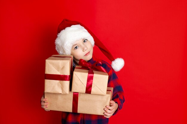 Bonnes vacances et joyeux Noël. Portrait d'un garçon dans une casquette avec des cadeaux dans ses mains sur fond rouge. Un endroit pour le texte. photo de haute qualité