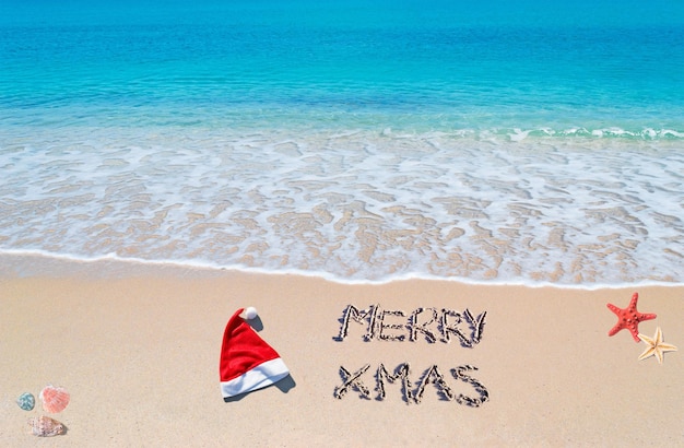 Photo bonnet de noel et joyeux noël écrit sur une plage tropicale