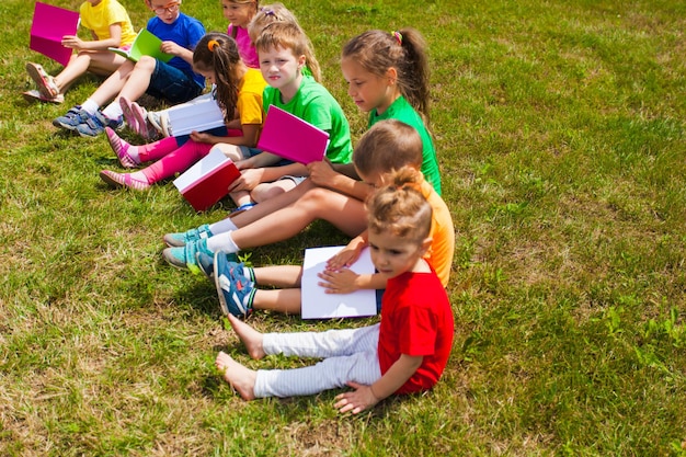 Les bons enfants en t-shirts colorés sont assis sur l'herbe avec des livres à la main