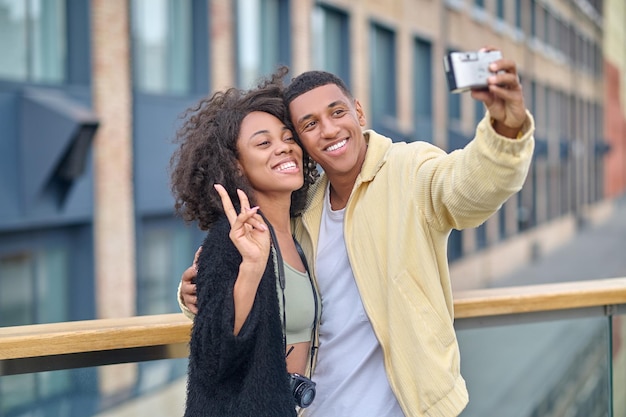 Bons moments. Un mec à la peau foncée étreignant une petite amie souriante montrant un signe de liberté avec la main prenant un selfie devant la caméra dans la rue de la ville