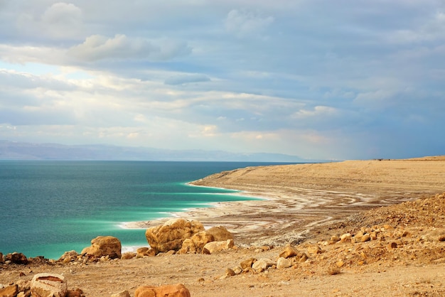 Bord de la mer Morte du côté de la Jordanie, plage de sable sec et de rochers, le soleil brille sur la belle surface de l'eau azur