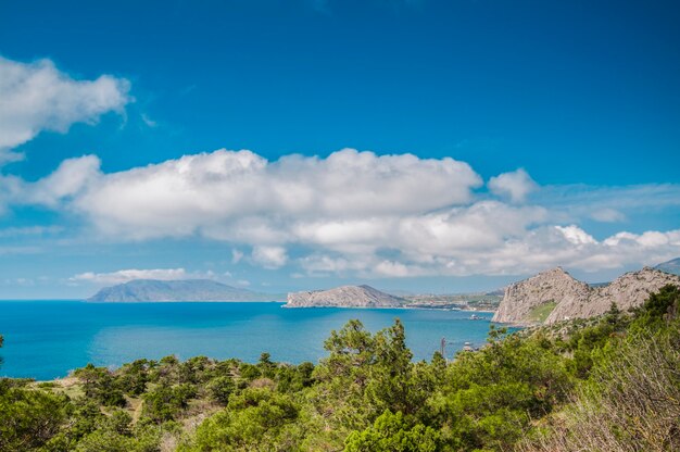 Bord de mer et plage rocheuse, ciel bleu avec nuages blancs, montagnes et petit village en arrière-plan