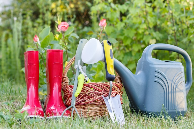 Bottes en caoutchouc de jardin rouge, petit râteau, sécateur, panier en osier, truelle et arrosoir en plastique sur l'herbe verte avec fond vert flou. Outils pour le jardinage