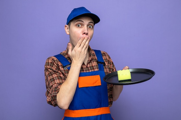 Bouche couverte concernée avec un jeune homme de nettoyage à la main portant un uniforme et une casquette tenant une éponge sur un plateau