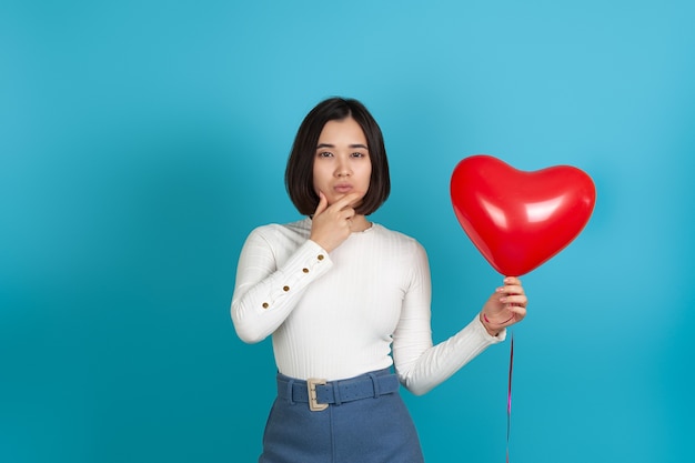 Bouchent la pensée pensive femme asiatique avec la main sous le menton, tenant un ballon en forme de coeur rouge