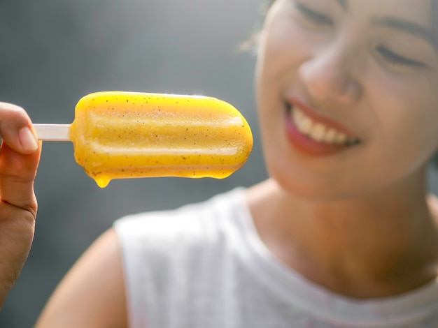 Bouchent le popsicle jaune dans la main d'une belle femme asiatique heureuse portant une chemise sans manches blanche décontractée, à l'extérieur. Femme mangeant des sucettes glacées. Femme souriante appréciant la sucette glacée en été.