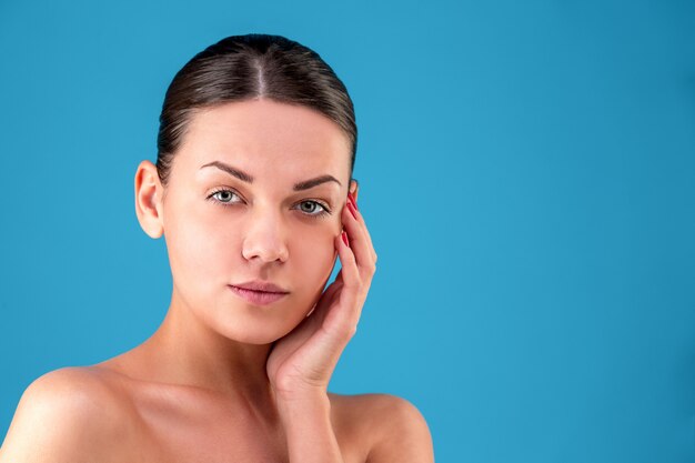 Bouchent le portrait de la beauté de la jeune femme brune souriante et touchant son visage sur l'espace bleu. Perfect Fresh Skin. Concept de jeunesse et de soins de la peau.