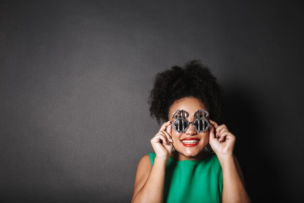 Bouchent le portrait d'une belle femme afro-américaine portant des lunettes de soleil en forme de dollar debout isolé sur un espace noir