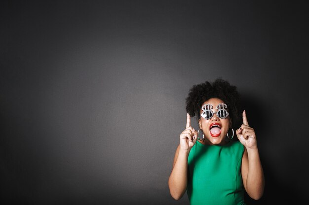 Bouchent le portrait d'une belle femme afro-américaine portant des lunettes de soleil en forme de dollar debout isolé sur un espace noir