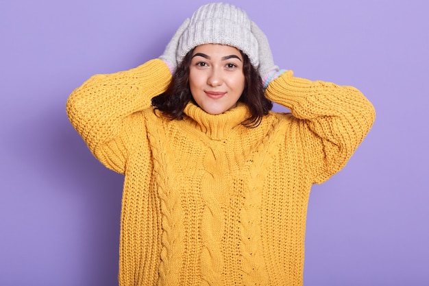 Bouchent portrait de charmante dame mince portant un chandail jaune et un bonnet blanc, regarde oyful, femme caucasienne posant avec un sourire sincère