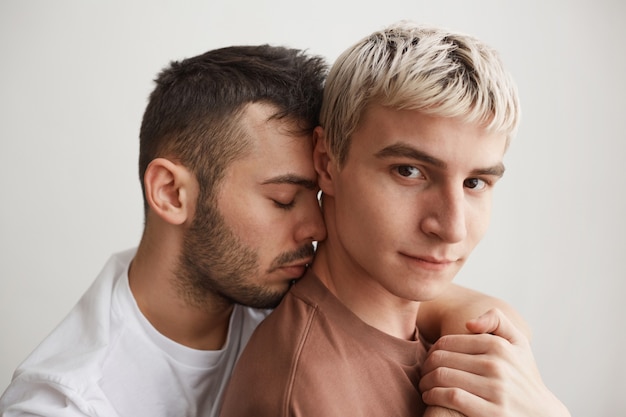 Photo bouchent le portrait d'un couple gay aimant embrassant et regardant la caméra en se tenant debout contre le blanc à la maison