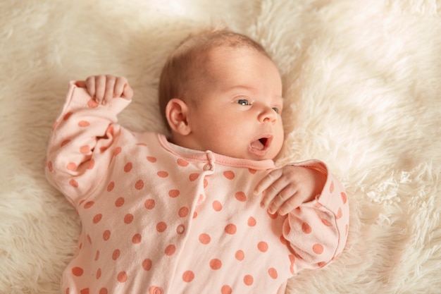 Bouchent le portrait du petit bébé couché dans son lit sur blanc moelleux, nouveau-né à la recherche de suite avec la bouche ouverte, charmant enfant portant un dormeur à pois.