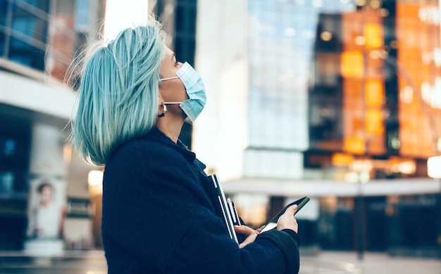 Bouchent le portrait d'une femme aux cheveux bleus portant un masque anti-grippe tout en tenant un ordinateur portable et son téléphone