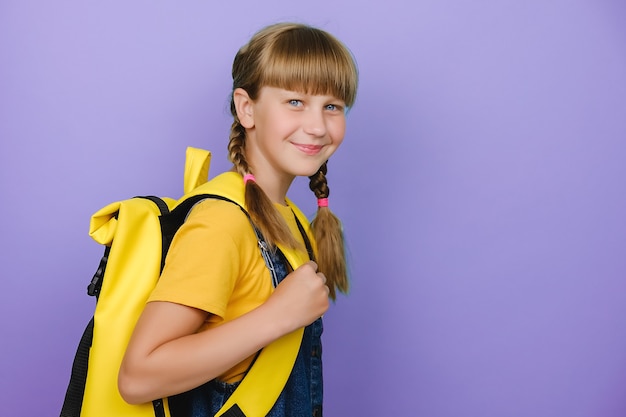 Bouchent le portrait d'une fille préadolescente souriante heureuse, écolière caucasienne tenant un sac à dos jaune debout isolée sur fond violet lilas, regardant la caméra de manière positive, concept de retour à l'école