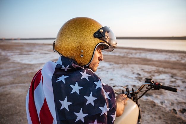 Bouchent le portrait d'un homme brutal portant un casque d'or et un drapeau américain assis sur la moto