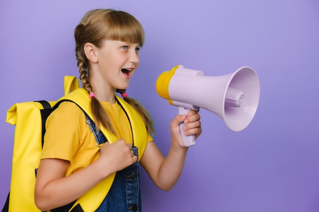 Bouchent le portrait d'une jeune adolescente blonde amusante à l'école avec un sac à dos jaune crier dans un mégaphone, posant isolée sur un studio d'enfants de mur de fond de couleur violette. Concept de mode de vie de l'éducation