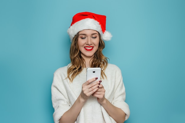 Bouchent le portrait de jeune femme en bonnet de Noel et chandail d'hiver blanc souriant et regardant dans le téléphone mobile