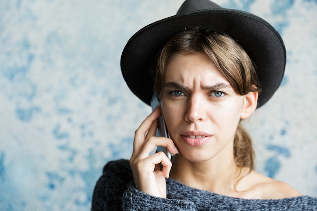 Bouchent Le Portrait D'une Jeune Femme Confuse Habillée En Chapeau Et Pull Parler Au Téléphone Mobile Sur Mur Bleu