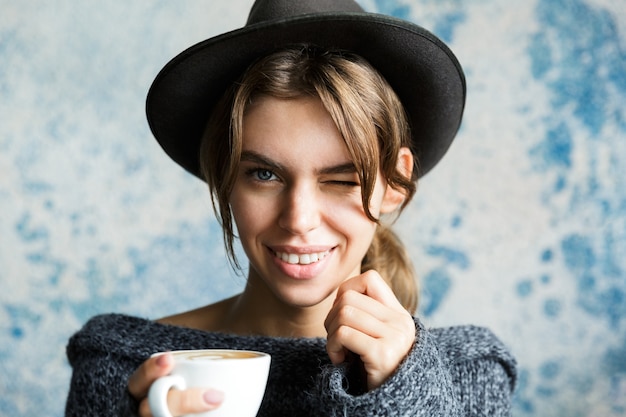 Bouchent le portrait d'une jeune femme habillée en pull et chapeau sur un mur bleu, tenant une tasse de café chaud, un clin de œil