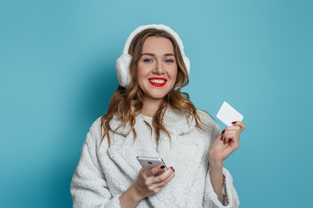 Bouchent le portrait de jeune femme souriante en manteau de fourrure blanche d'hiver tenant un téléphone mobile et une carte bancaire de crédit isolée sur mur bleu