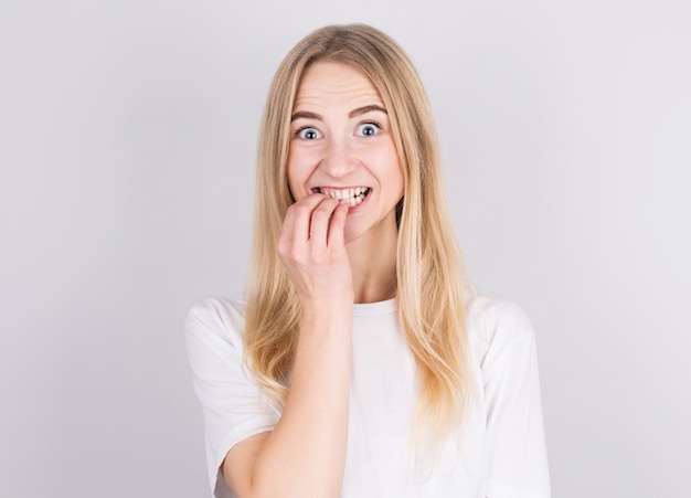 Bouchent le portrait d'une jeune fille blonde inquiète en t-shirt blanc se mordant les ongles isolés