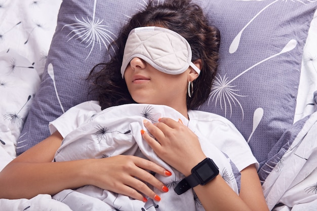 Bouchent le portrait de jeune fille avec un masque pour les yeux dormant sous une couverture, ayant un travail acharné et veut se détendre, allongé dans son lit sur un oreiller, ayant les cheveux noirs, un t-shirt blanc habillé, a une horloge noire à portée de main.