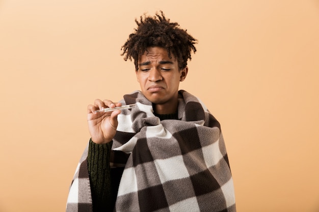 Photo bouchent le portrait d'un jeune homme africain bouleversé enveloppé dans une couverture isolée sur un mur beige, montrant un thermomètre