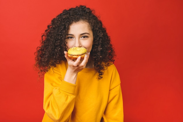 Bouchent Le Portrait D'une Jolie Jeune Fille Satisfaite De Manger Des Beignets Isolés Sur Fond Rouge.