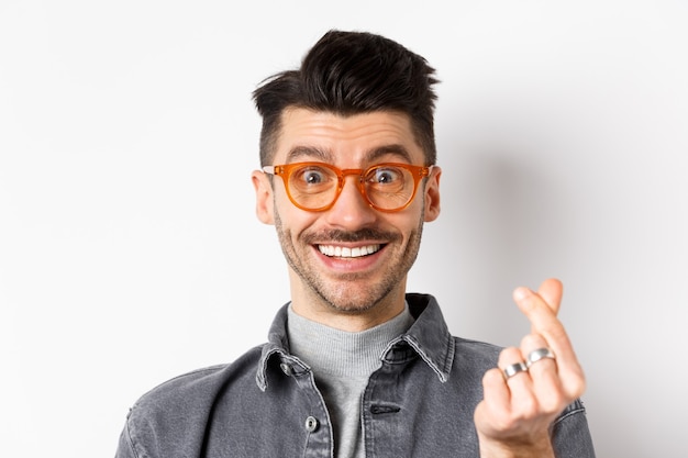 Bouchent le portrait d'un mec mignon avec une moustache portant des lunettes, montrant le signe du cœur de la main, souriant heureux, debout sur fond blanc.