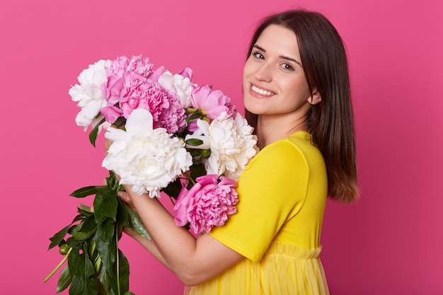 Bouchent le portrait de profil de jolie femme élégante en t-shirt décontracté jaune isolé sur studio rose, tenant le bouquet de fleurs de pivoine, femme avec une expression faciale heureuse. Concept d'amour.