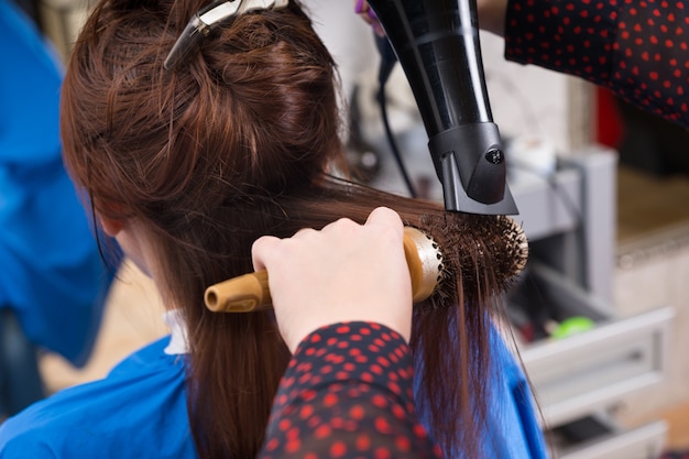 Photo bouchent la vue arrière d'une femme brune ayant les cheveux mouillés, brossés et séchés par un styliste à l'aide d'une brosse ronde et d'un sèche-cheveux à main dans le salon