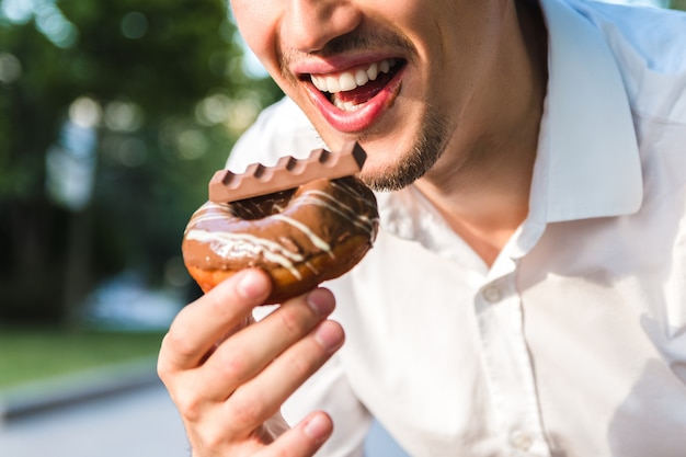 Bouchent la vue du beau jeune homme caucasien en chemise blanche bénéficiant d'un délicieux beignet au chocolat dans le parc de la ville. nourriture indésirable mais savoureuse pour une humeur positive.