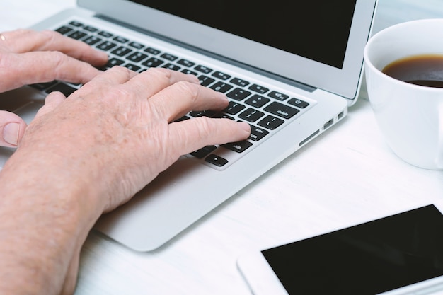 Bouchent la vue des mains senior sur le clavier avec une tasse de café un téléphone.