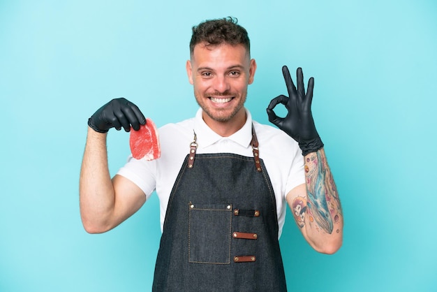 Boucher homme caucasien portant un tablier et servant de la viande fraîchement coupée isolée sur fond bleu montrant un signe ok avec les doigts