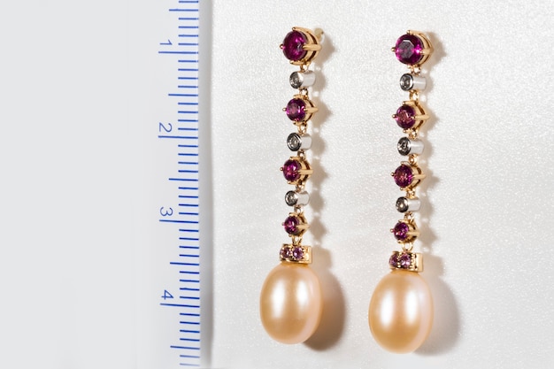 Boucles d'oreilles longues en or avec diamants, rubis et perles, sur fond blanc à côté de la règle