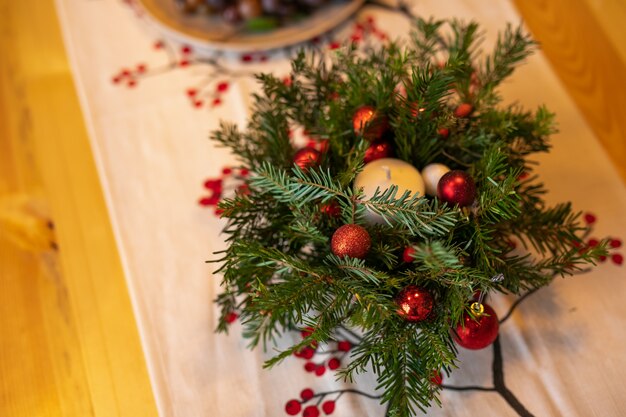 Une bougie blanche décorée de branches de sapin et de boules de verre rouge debout sur une table en bois avec une serviette en lin.