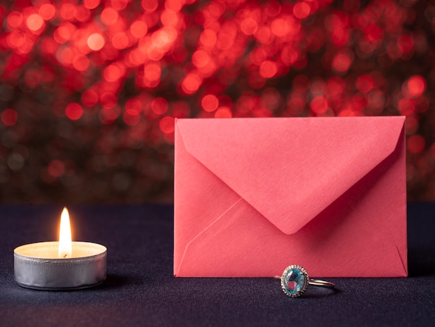 Une bougie blanche qui brûle et à côté se trouve une carte rose devant laquelle un anneau avec une pierre bleue, sur un rose