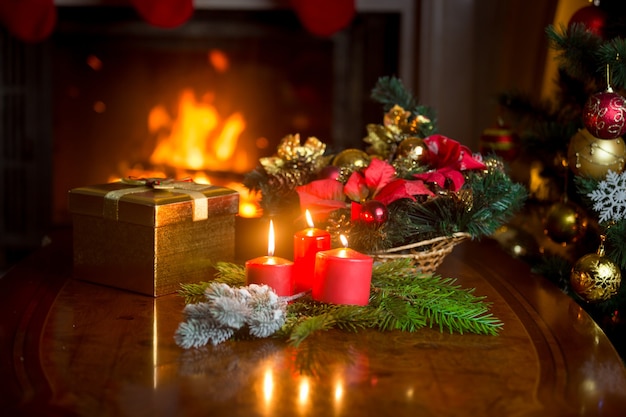 Bougies allumées, couronne de Noël et coffret cadeau doré sur la table à côté d'une cheminée allumée dans le salon. Image avec une faible profondeur de champ.