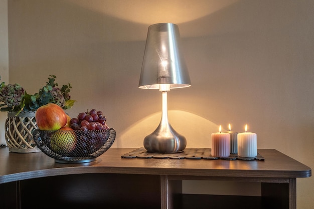 Des bougies allumées et un vase avec des pommes de fruits et des raisins sur la table près de la lampe