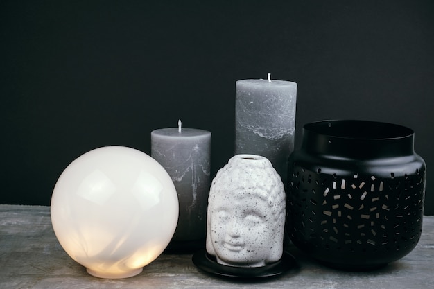 Bougies grises, lampe ronde blanche et tête de Bouddha en céramique blanche sur une table en face du mur noir