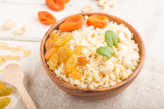 Bouillie de boulgour aux abricots secs, raisins secs et noix de cajou dans un bol en bois sur une surface en bois blanc et textile en lin. Vue de côté, gros plan, mise au point sélective