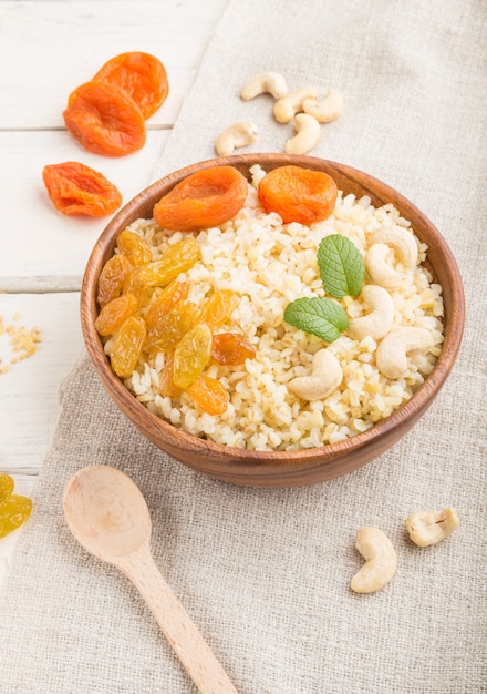 Bouillie de boulgour aux abricots secs, raisins secs et noix de cajou dans un bol en bois sur une surface en bois blanc et textile en lin. Vue latérale, gros plan.