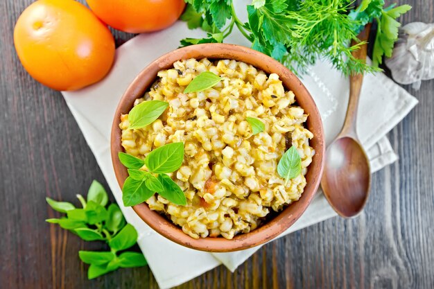 Bouillie d'orge dans un bol en argile avec du basilic sur serviette, tomates, persil et ail sur fond de planches de bois sur le dessus